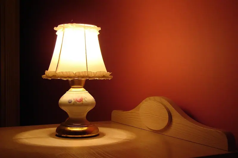 Abajur de cúpula clara aceso sobre uma mesa de cabeceira, junto à cabeceira de madeira da cama