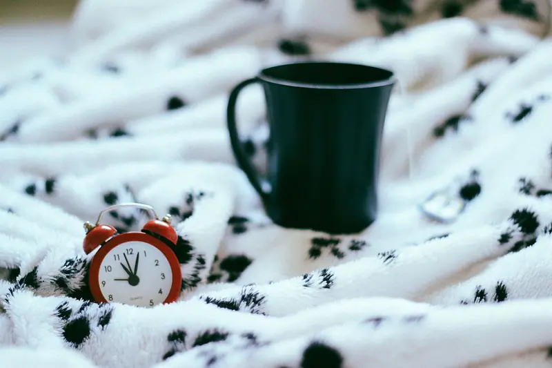 Pequeno despertador vermelho e caneca verde sobre uma manta branca com estampa preta