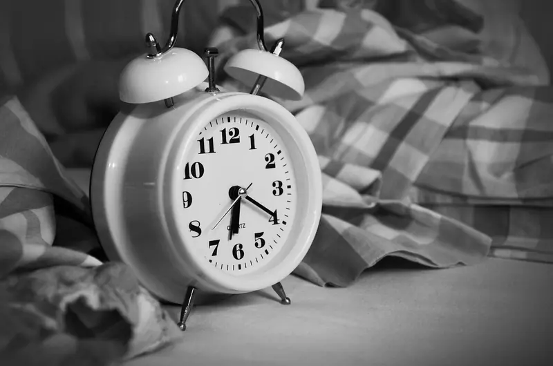 Despertador branco sobre uma cama desarrumada com cobertor xadrez, em preto e branco