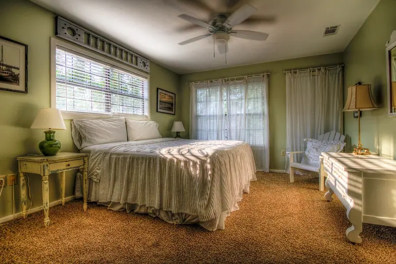 Quarto com cama arrumada, abajures, poltrona branca e janelas amplas com cortinas claras
