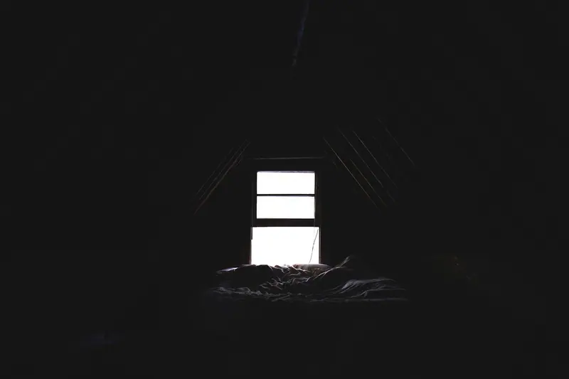 Quarto escuro sob um telhado inclinado, com uma pequena janela iluminada acima da cama