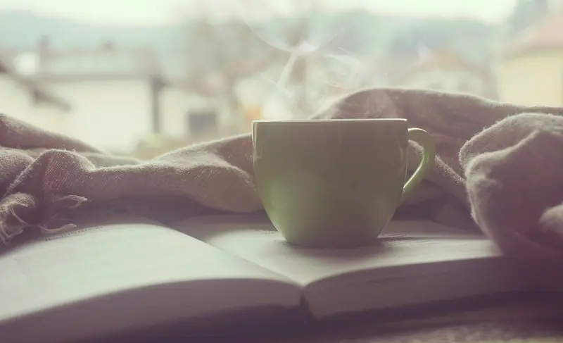 Xícara verde sobre um livro aberto e uma manta, perto de uma janela com luz suave