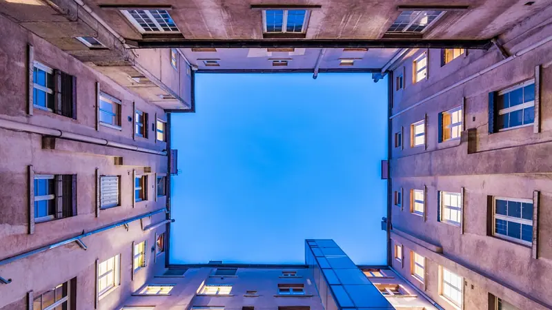 Pátio interno de um prédio visto de baixo, com janelas em volta e o céu azul no alto