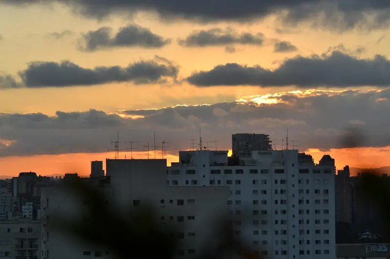 Céu alaranjado do fim de tarde atrás de prédios residenciais, com galhos em primeiro plano