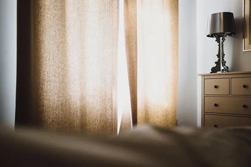Quarto à meia-luz com cortina clara iluminada pelo sol e um abajur sobre a cômoda