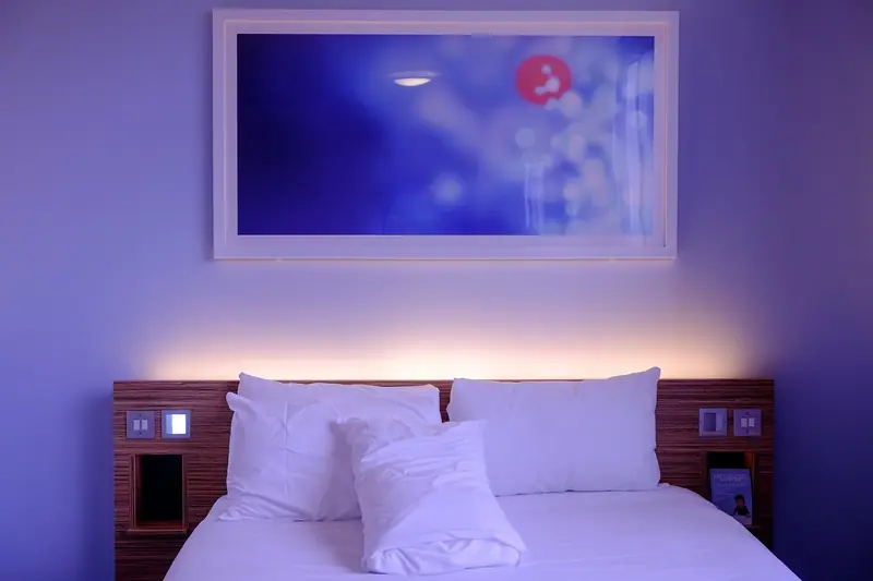 Cama de casal com travesseiros brancos num quarto à meia-luz, com luz azulada