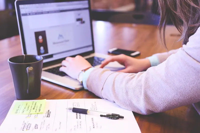 Mãos digitando no notebook sobre uma mesa de madeira, com caneca e anotações ao lado
