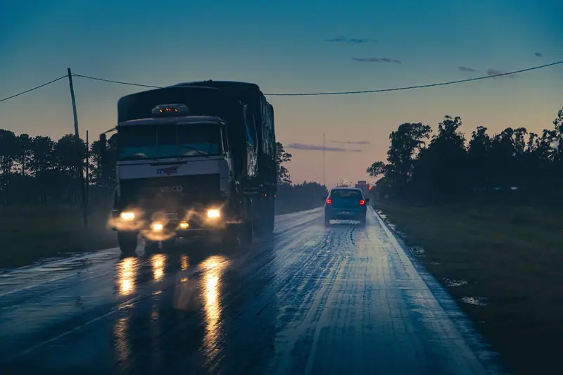 Caminhão com faróis acesos numa estrada molhada ao entardecer