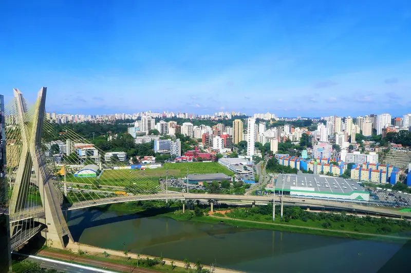 Rio com ponte estaiada e avenidas nas margens, visto do alto, com prédios e bairros até o horizonte