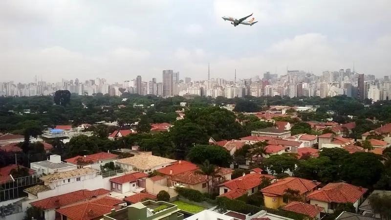 Avião cruzando o céu sobre casas de telhado vermelho e prédios de São Paulo