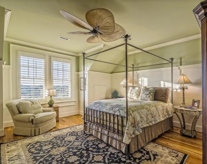 Quarto com cama de dossel, poltrona, tapete e janelas com venezianas brancas