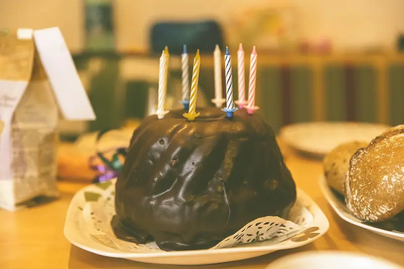 Bolo de chocolate com velinhas coloridas ainda apagadas, sobre uma mesa com pães e pratos