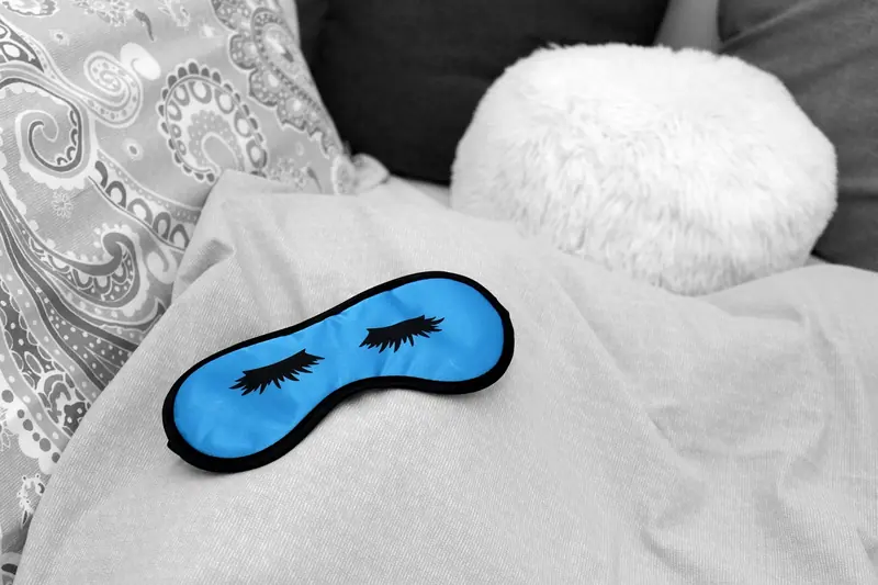 Máscara de dormir azul sobre lençóis cinza, ao lado de uma almofada branca