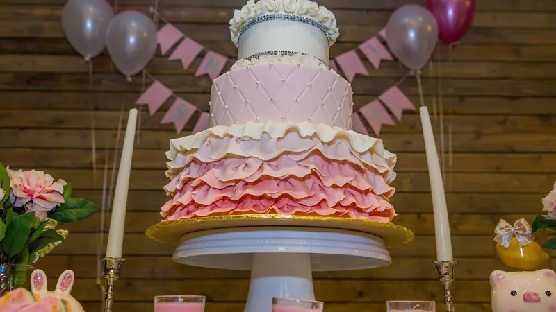 Mesa de festa com bolo rosa de três andares, velas, bandeirinhas e balões presos numa parede de madeira