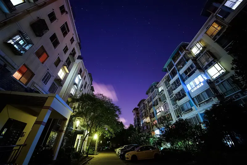 Rua entre prédios residenciais à noite, com janelas acesas e carros estacionados junto à calçada