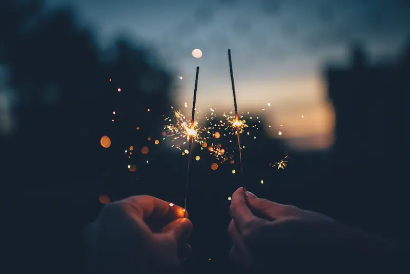 Mãos segurando duas estrelinhas acesas ao entardecer, com faíscas douradas