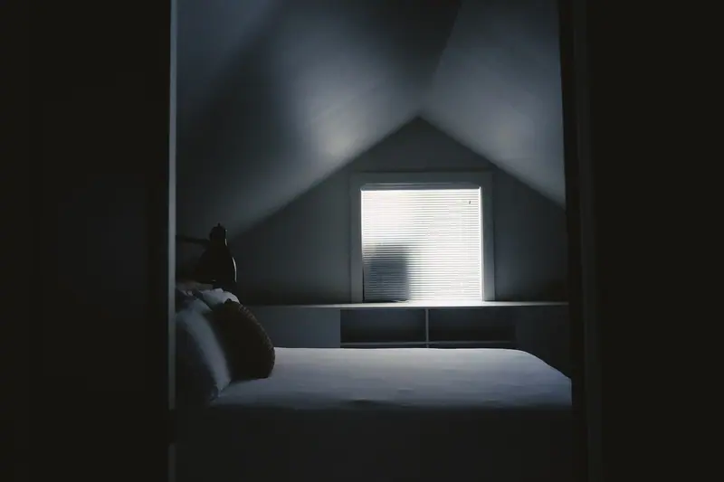 Quarto escuro com uma cama e a claridade de uma janela ao fundo