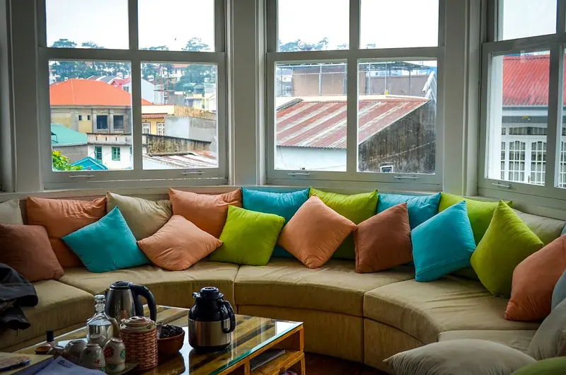 Sala com sofá curvo cheio de almofadas coloridas e janelas voltadas para telhados vizinhos