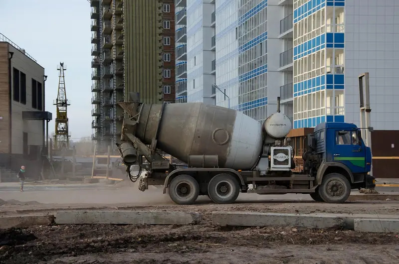 Caminhão betoneira num canteiro de obras de terra batida, ao lado de prédios residenciais
