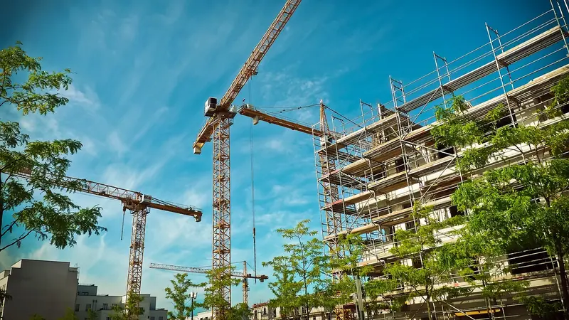 Guindastes de obra e um prédio coberto de andaimes sob céu azul, com árvores em primeiro plano
