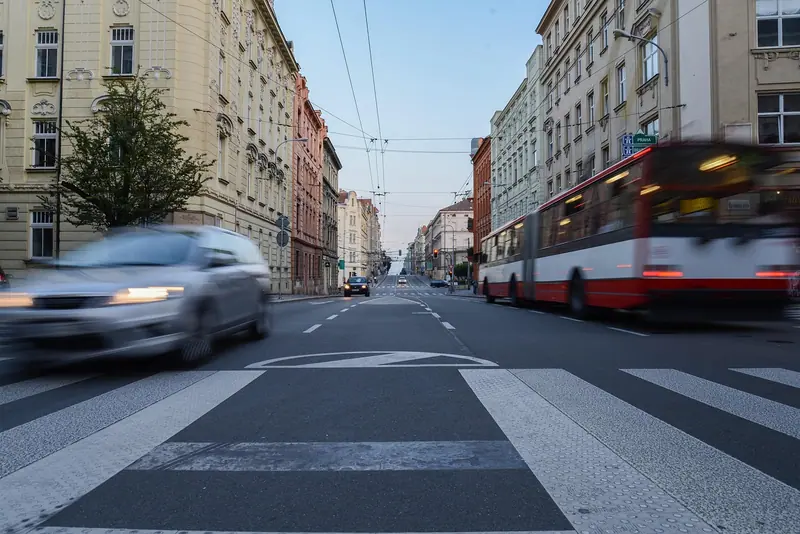Ônibus articulado vindo pela avenida, com um carro passando rápido perto da faixa de pedestres
