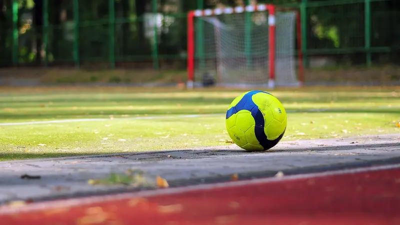 Bola amarela e azul parada numa quadra de grama sintética, com um gol pequeno e alambrado verde ao fundo