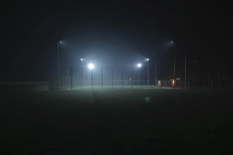 Campo esportivo escuro, com refletores acesos numa noite de neblina