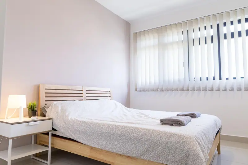 Quarto claro com cama de madeira, abajur aceso e janela com persiana vertical