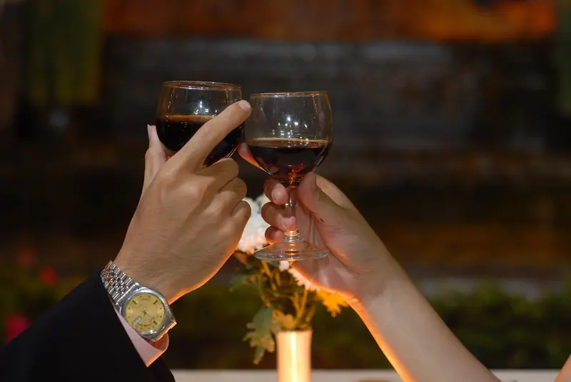 Duas mãos brindando com taças de vinho tinto, com um arranjo de flores ao fundo