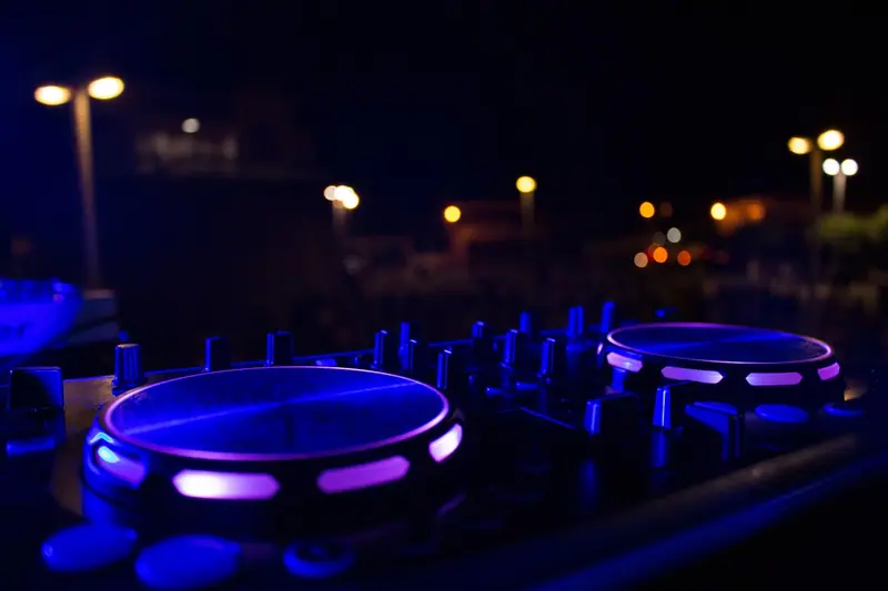 Controladora de DJ iluminada em azul e roxo, ao ar livre, com postes acesos ao fundo