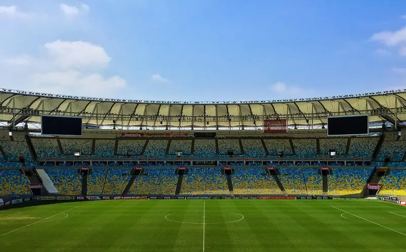 Estádio de futebol vazio num dia de céu azul, com arquibancadas e gramado