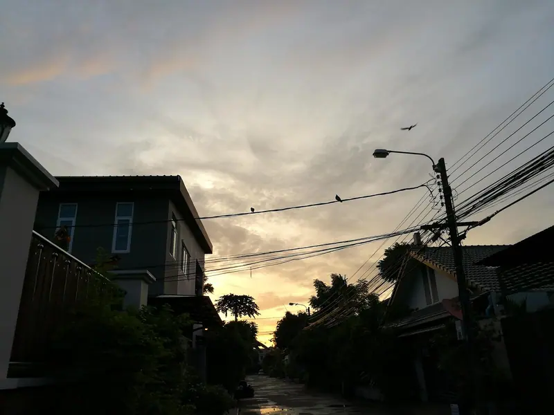Rua residencial ao entardecer, com casas dos dois lados, um poste de luz e muitos fios elétricos