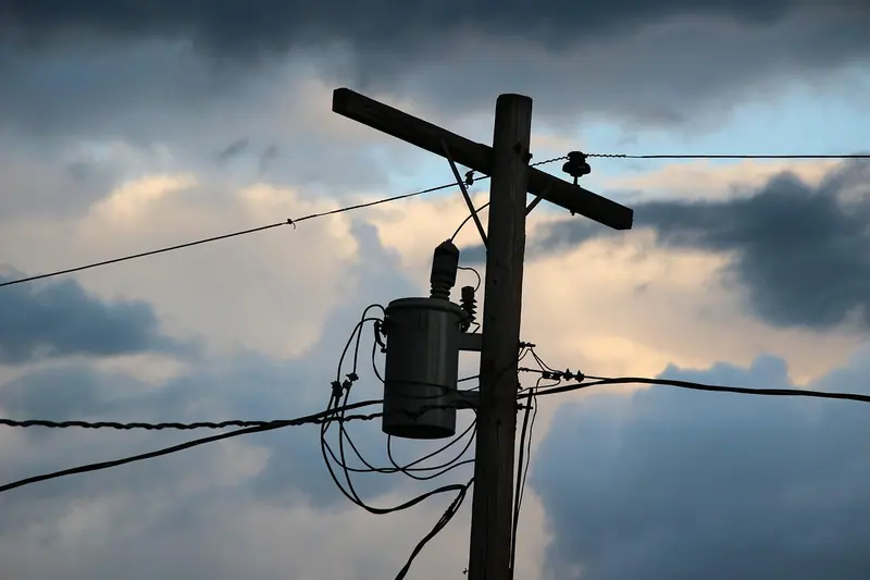 Poste de madeira com um transformador cilíndrico e vários fios, sob um céu nublado de fim de tarde