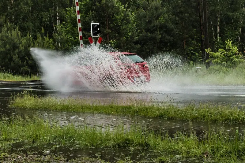 Carro vermelho passando por uma poça grande e levantando um leque de água ao lado da pista