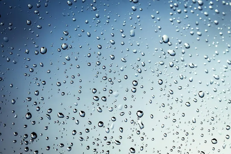 Gotas de chuva de vários tamanhos espalhadas num vidro, com o céu azulado ao fundo