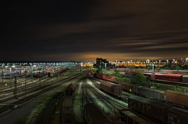 Pátio ferroviário à noite, com muitos vagões de carga parados sobre trilhos e luzes da cidade no horizonte