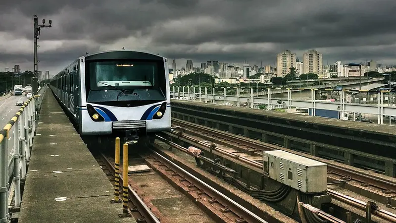 Trem de metrô chegando por um trecho a céu aberto, com prédios ao fundo e céu nublado