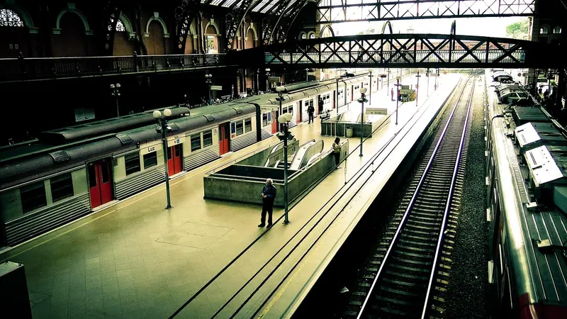 Plataforma de estação com um trem parado ao lado dos trilhos e passageiros ao longe