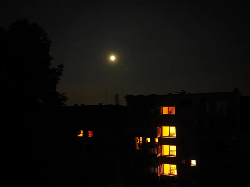 Prédio com algumas janelas acesas à noite, com a lua no céu escuro
