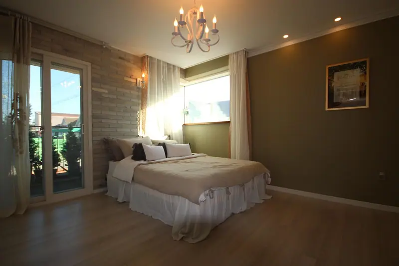 Quarto com cama de casal, cortinas claras e luz do dia entrando pela janela