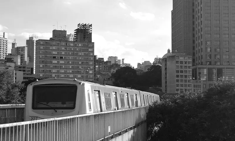 Trem de metrô passando por um viaduto entre prédios residenciais, em foto preto e branco