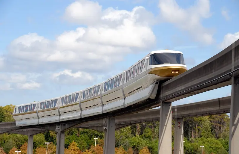 Trem de monotrilho branco correndo sobre uma viga elevada de concreto, com árvores e céu azul ao fundo