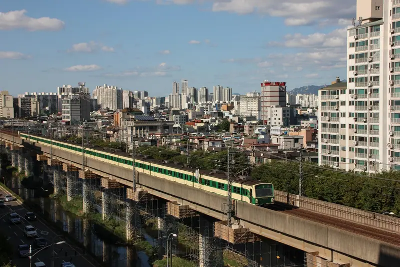 Trem passando por um viaduto ao lado de prédios residenciais altos, com uma avenida movimentada embaixo