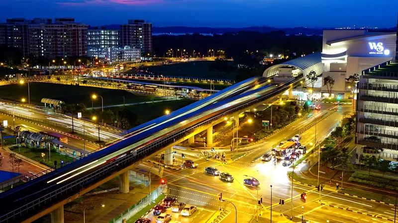 Via elevada de trem cruzando uma avenida à noite, com o rastro de luz de um trem e carros iluminados embaixo