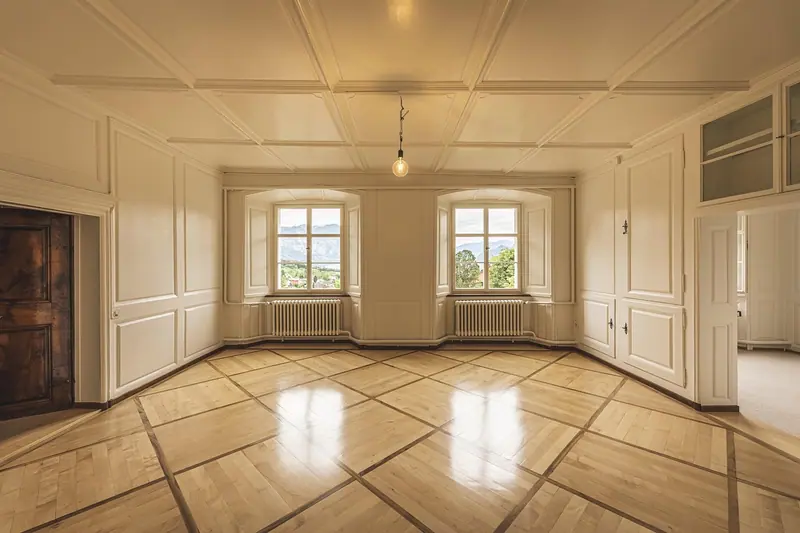 Cômodo vazio com piso de madeira encerado, paredes claras e duas janelas ao fundo