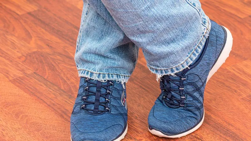 Pés de uma pessoa de tênis e calça jeans em pé sobre um piso de madeira