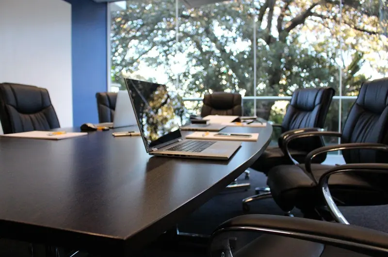 Sala de reunião com mesa escura, cadeiras de couro, notebook e paredes de vidro com vista para árvores