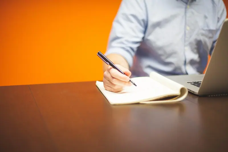 Mão escrevendo num caderno sobre a mesa, ao lado de um notebook, diante de uma parede laranja