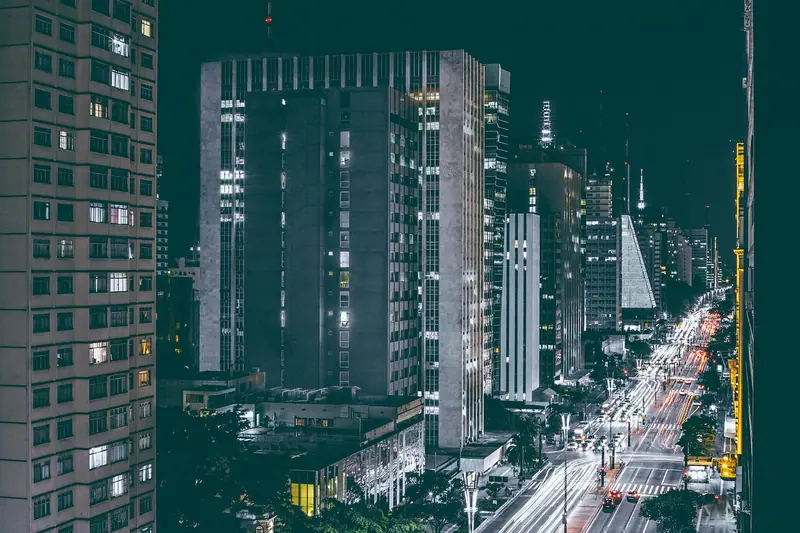 Avenida movimentada entre prédios altos à noite, com rastros de luz dos carros