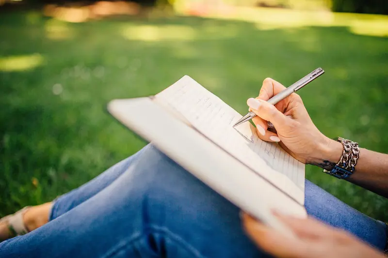Mãos escrevendo num caderno apoiado sobre as pernas, ao ar livre
